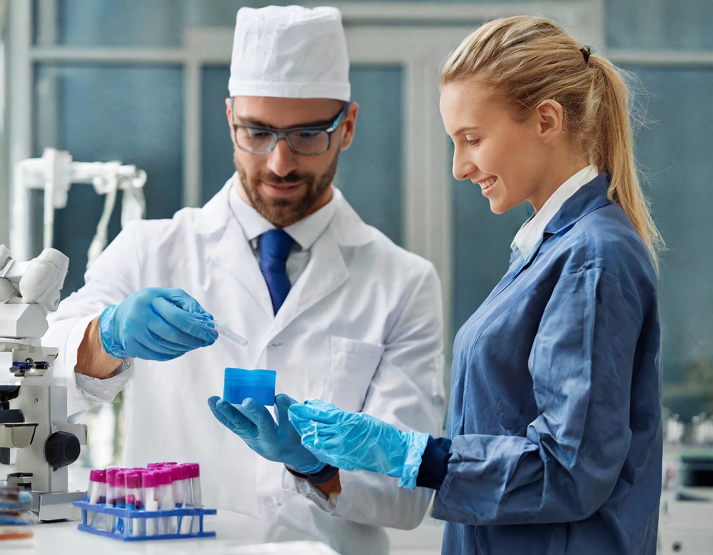 The image shows a man holding a plastic-molded part and a woman standing beside him. Both wear gowns in a medical environment.