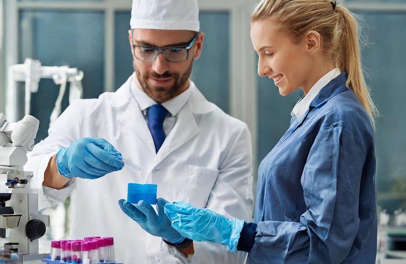 The image shows a man holding a plastic-molded part and a woman standing beside him. Both wear gowns in a medical environment.